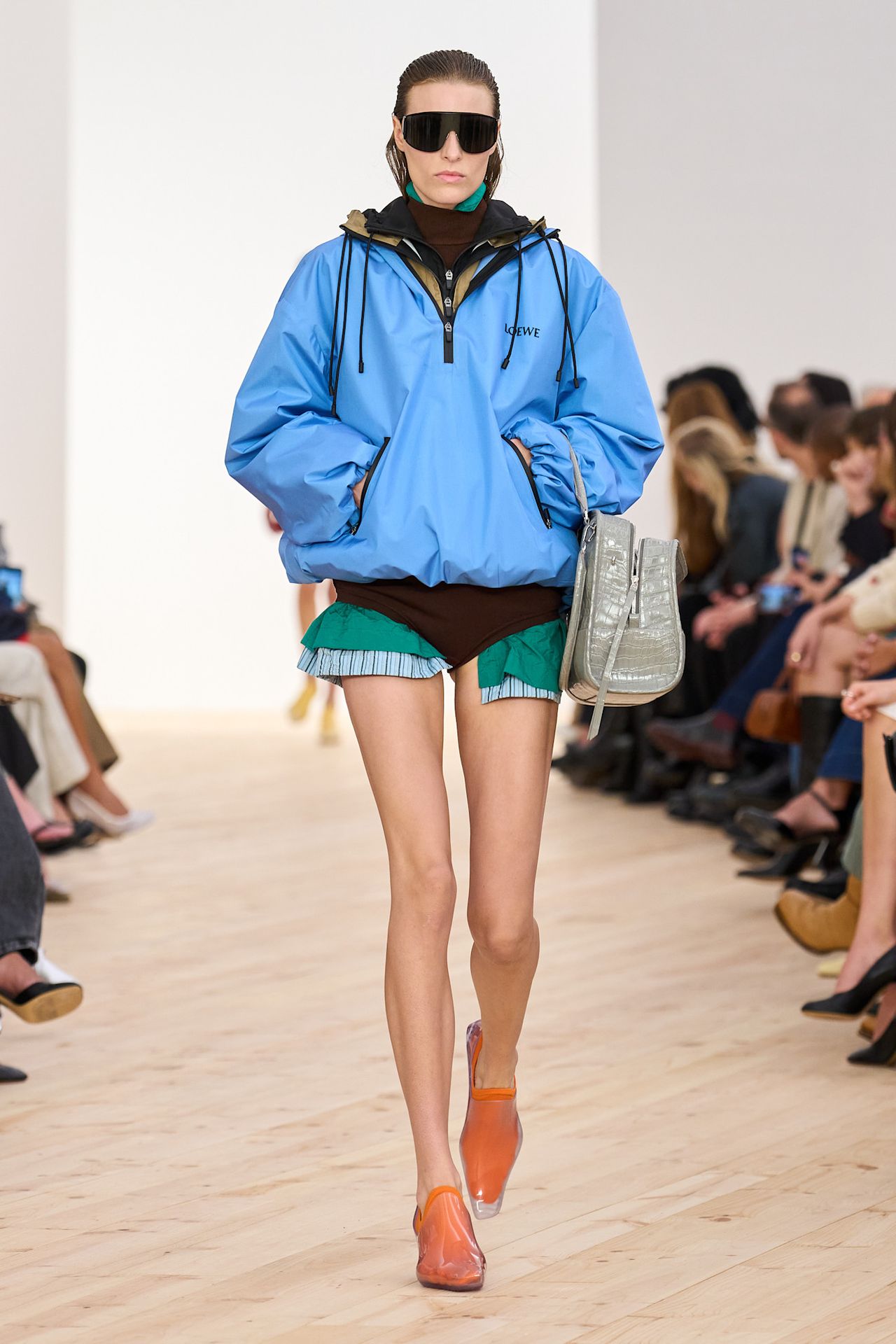 Layering staples for spring 2026 is shown in a photo from Loewe's runway show, with a model walking while wearing a bright blue parka layered over a tan parka jacket, a dark brown turtleneck bodysuit, and a bright green button-down and a blue striped button-down. The rest of the look is styled with oversized aviators, a gray crocodile bag, and clear pointed loafers with bright orange socks