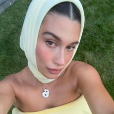 Hailey Bieber wearing a yellow Jacquemus headscarf with her B necklace and a matching Jacquemus fall 2024 strapless dress in a selfie.
