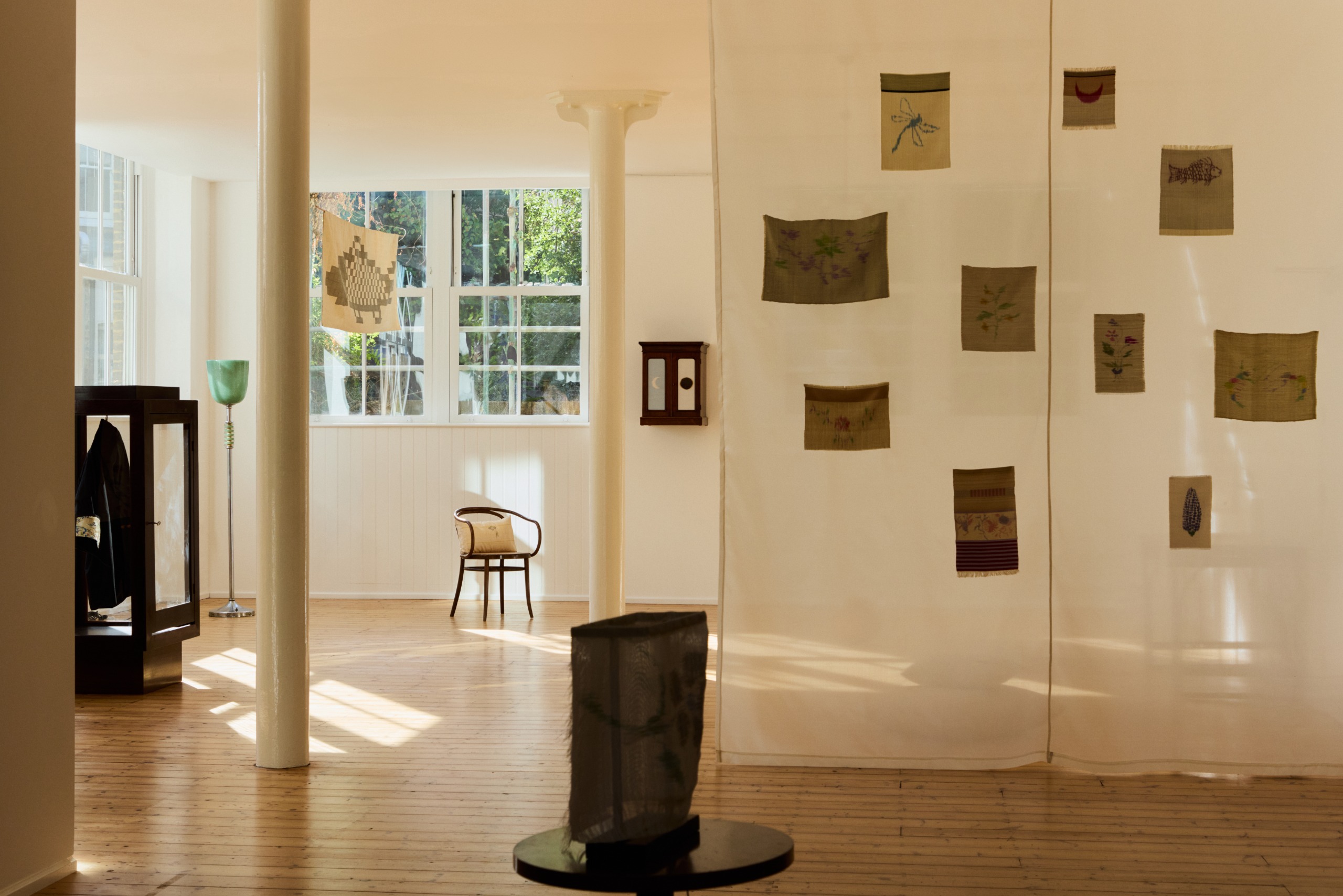 A series of woven textile creations in large and small formats hang in a sun-lit gallery with wooden floors and curated objects installed throughout it.