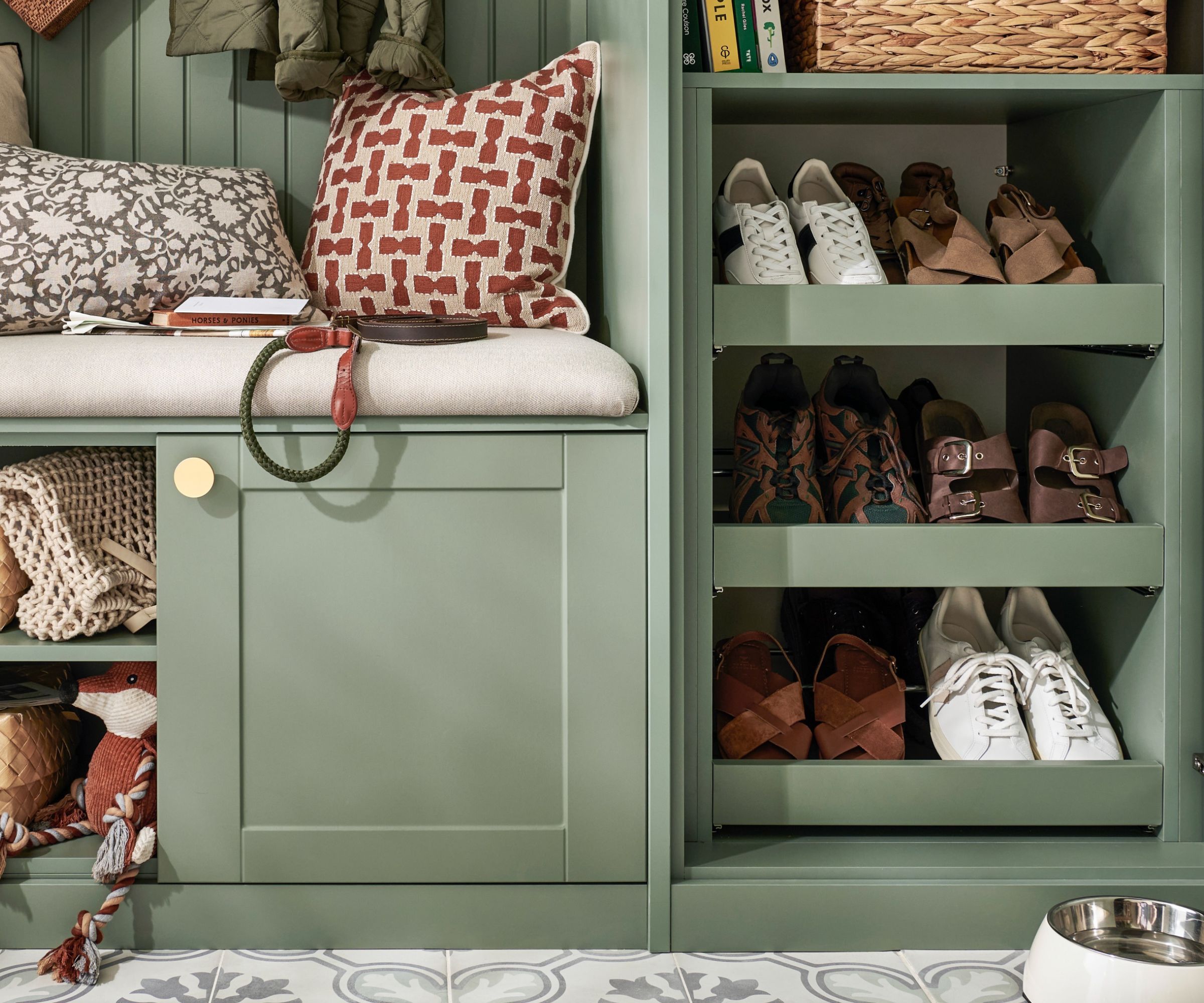A sage green painted built-in hallway unit with a cushioned bench seat, brass knob door and open cubbies below, alongside a tall pull-out shoe storage tower with three angled shelves holding trainers.