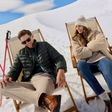 Man and woman sat on loungers in the snow wearing Crew Clothing Winter outerwear