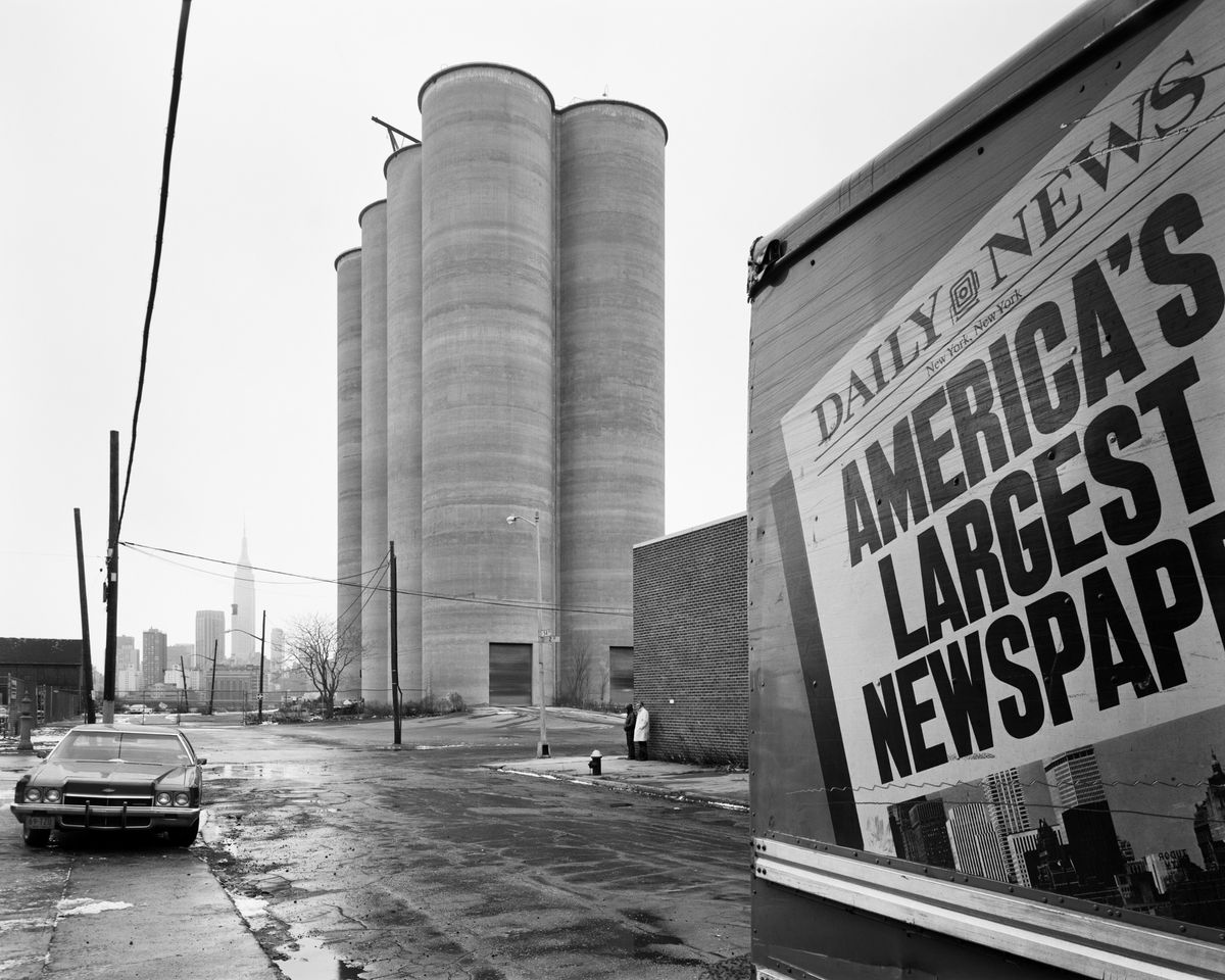 A parked car and a truck bearing a Daily News advertisement sit near a row of tall concrete grain silos, with the Empire State Building visible in the distance.