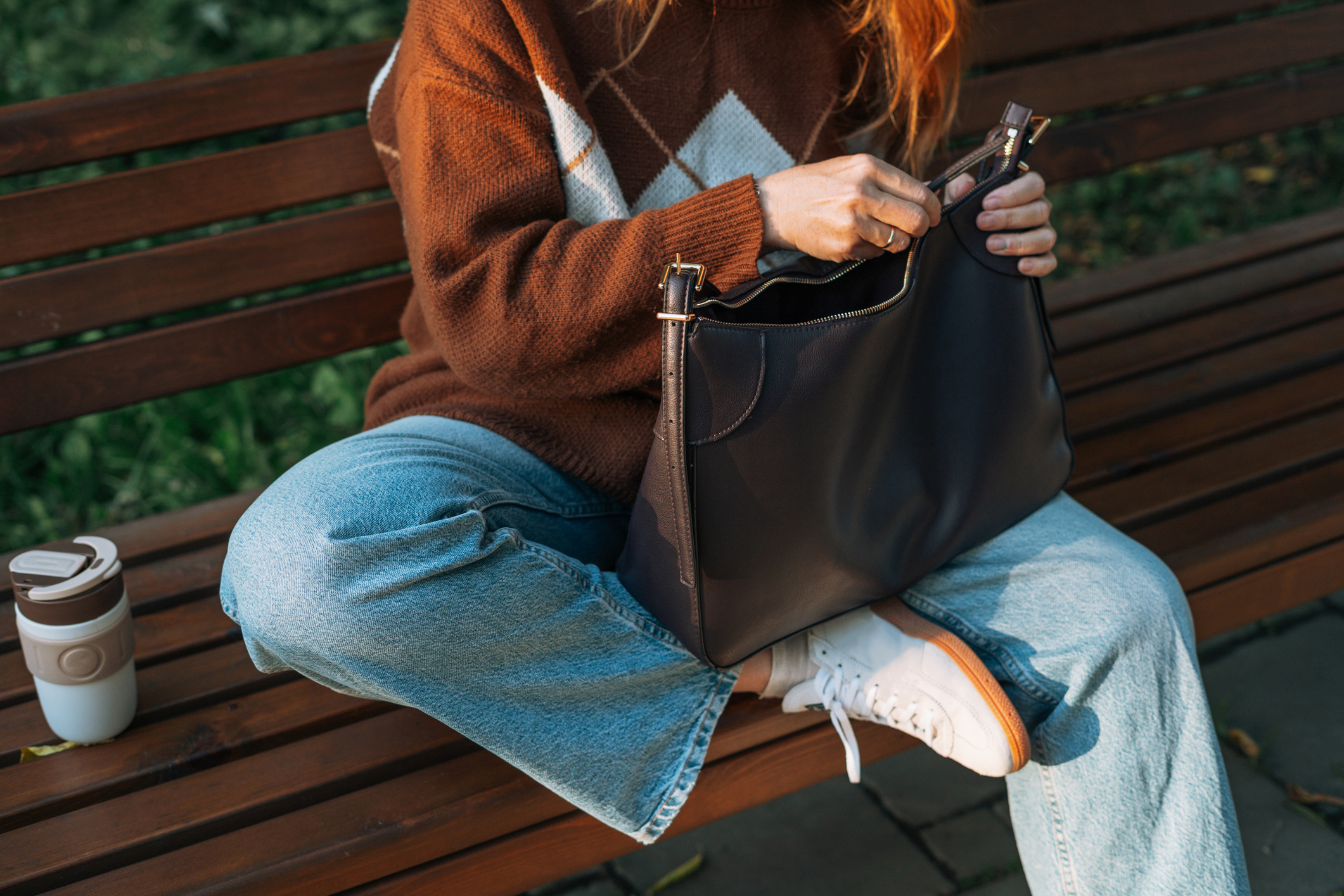 A woman sat on a bench wearing a brown jumper and jeans, zipping up her brown tote bag.