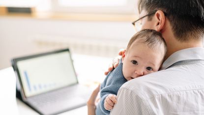 A man holds his new baby on his shoulder while he looks at his tablet at his desk at home.