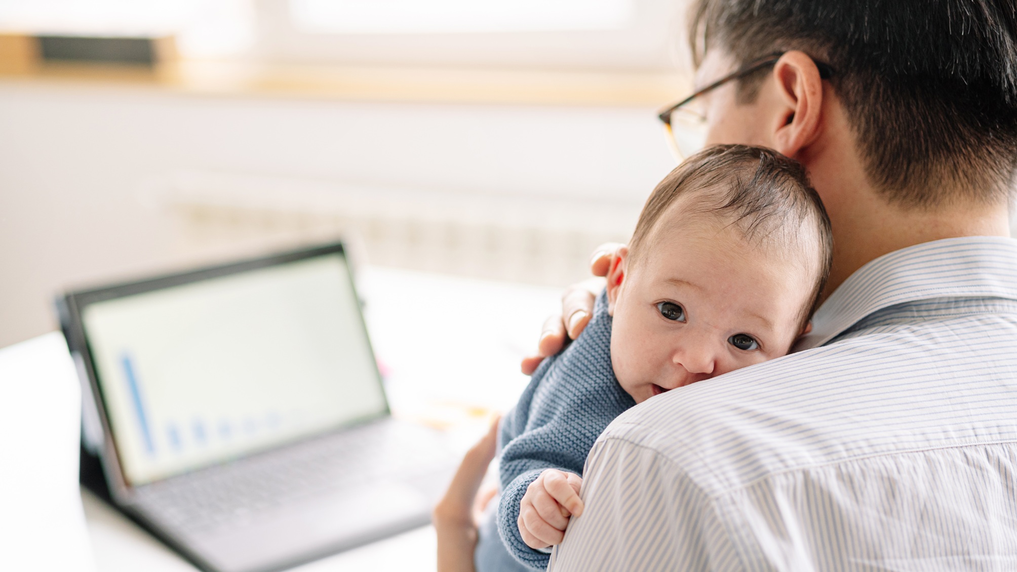 A man holds his new baby on his shoulder while he looks at his tablet at his desk at home.