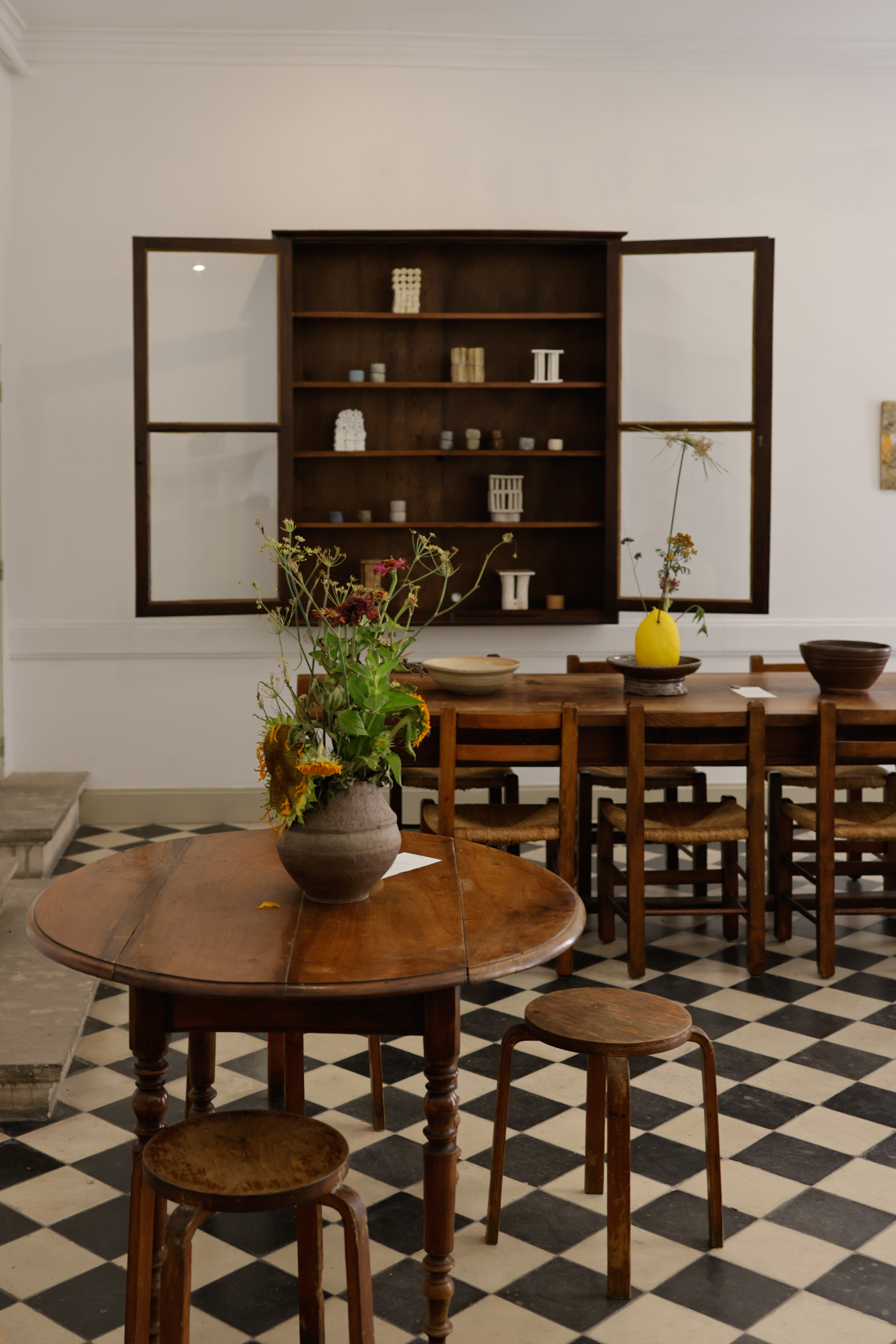 A gallery and coffee shop features checkered black and white floors, vintage wooden furniture, organic ceramics, and a glass and wood vitrine filled with collectibles.