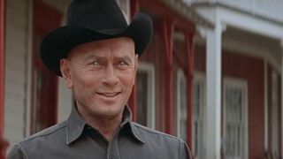 a clean-shaven man with a black cowboy-style hat looks past the camera with light reflecting in his eyes