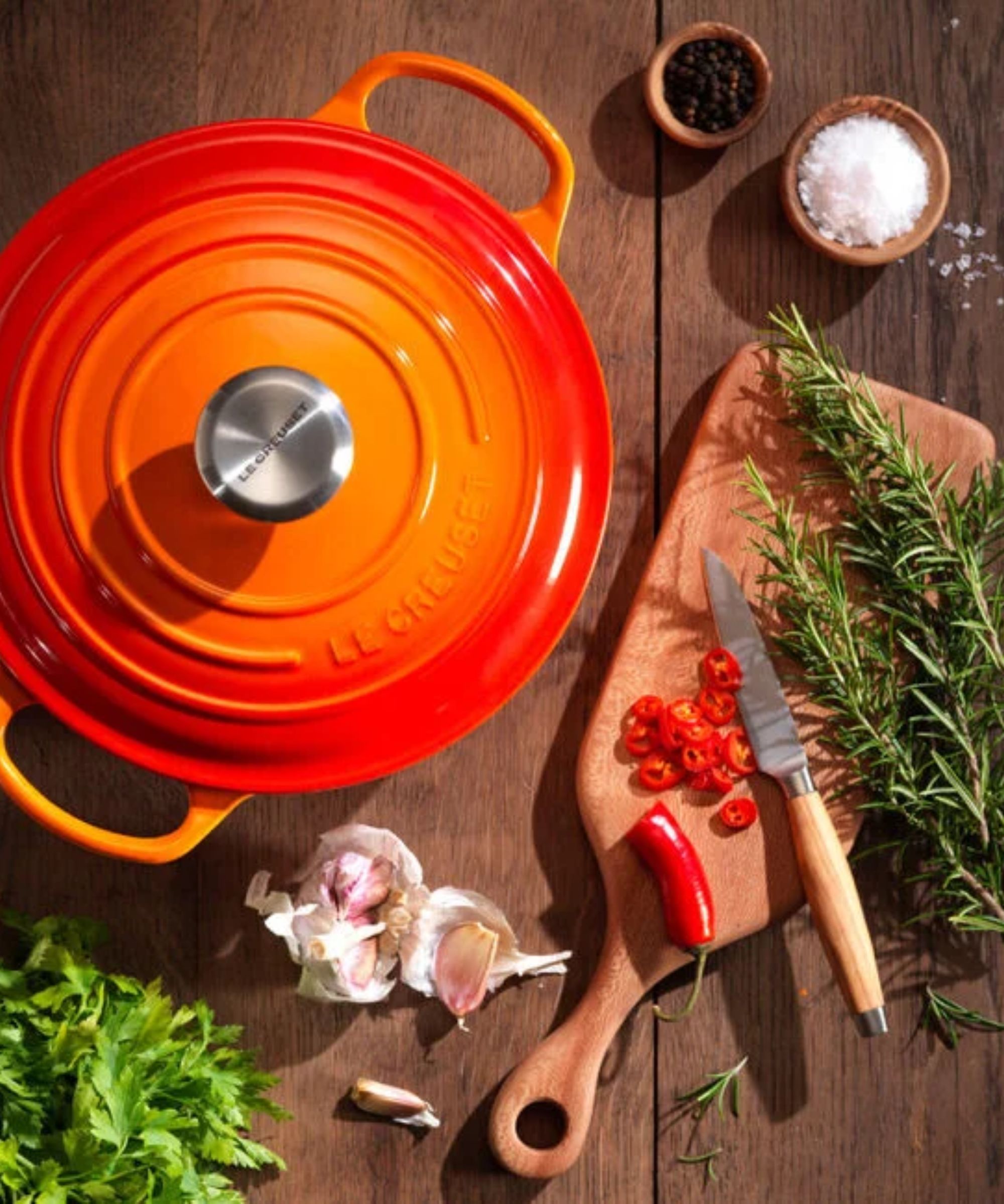 A Red Le Creuset Dutch Oven on a table surrounded by fresh herbs and chillies.