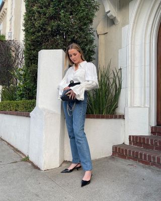 Fashion editor Kristen Nichols poses outside a building in Los Angeles wearing a white button-down with statement sleeves, straight-leg jeans, and black slingback kitten heels.