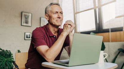 An older man sits in front of his laptop at his desk at home.