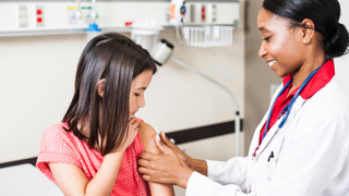 A girl is shown looking at her arm as a doctor places a band aid on it after having a vaccine