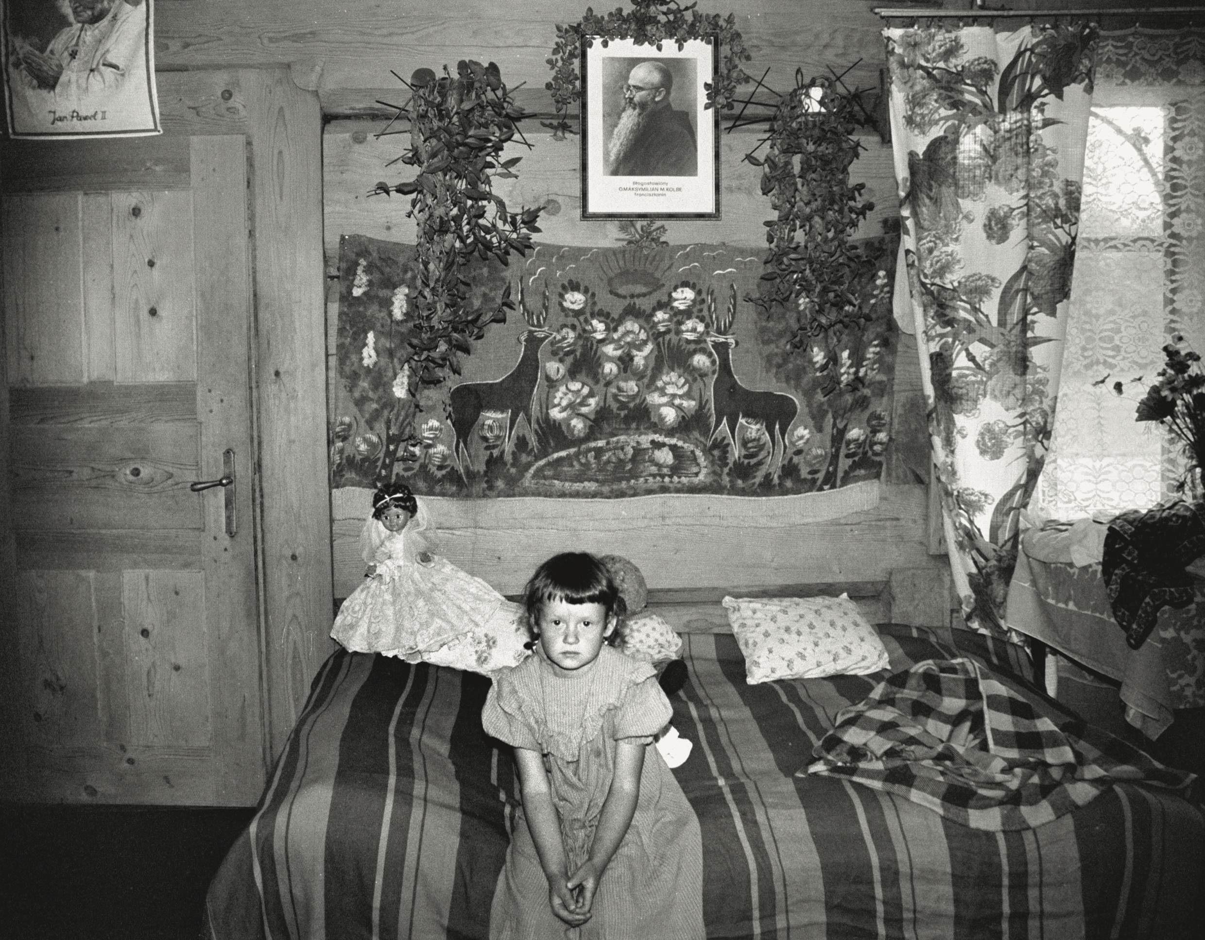 Zofia Rydet black and white photograph of a girl sat on the edge of a bed in Poland. A toy doll can be seen behind her.