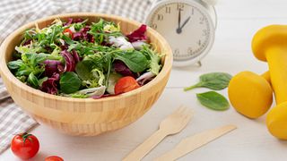 Clock on table with bowl of salad and weights to symbolise intermittent fasting