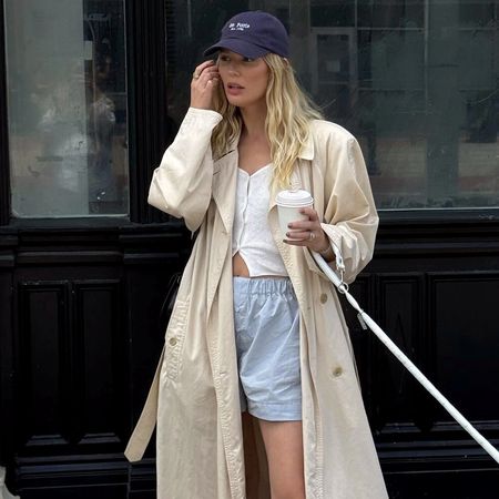 style influencer Megan Adelaide Vega poses while walking her dog in a navy baseball cap, long trench coat, white button-down knit top, and blue boxer shorts