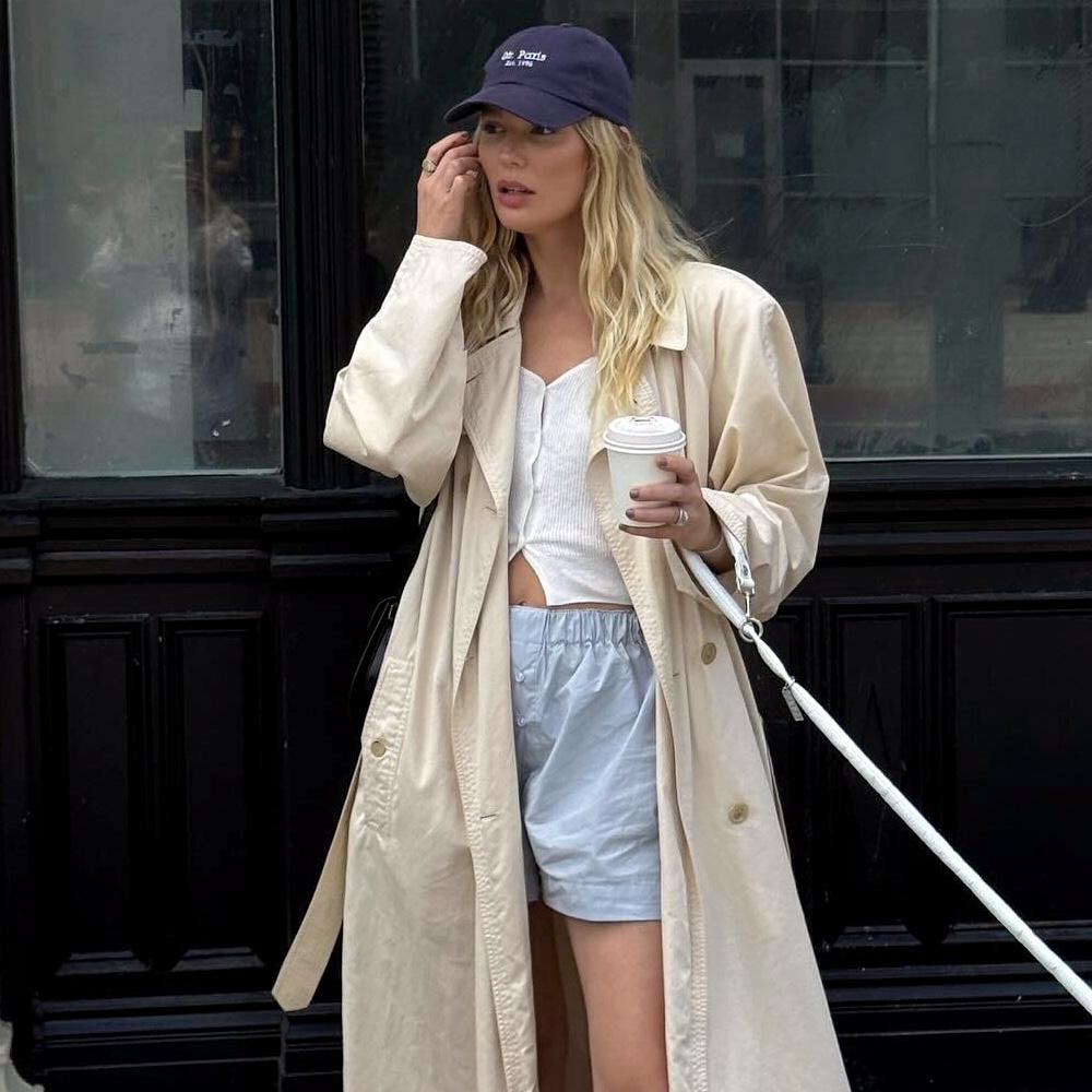style influencer Megan Adelaide Vega poses while walking her dog in a navy baseball cap, long trench coat, white button-down knit top, and blue boxer shorts