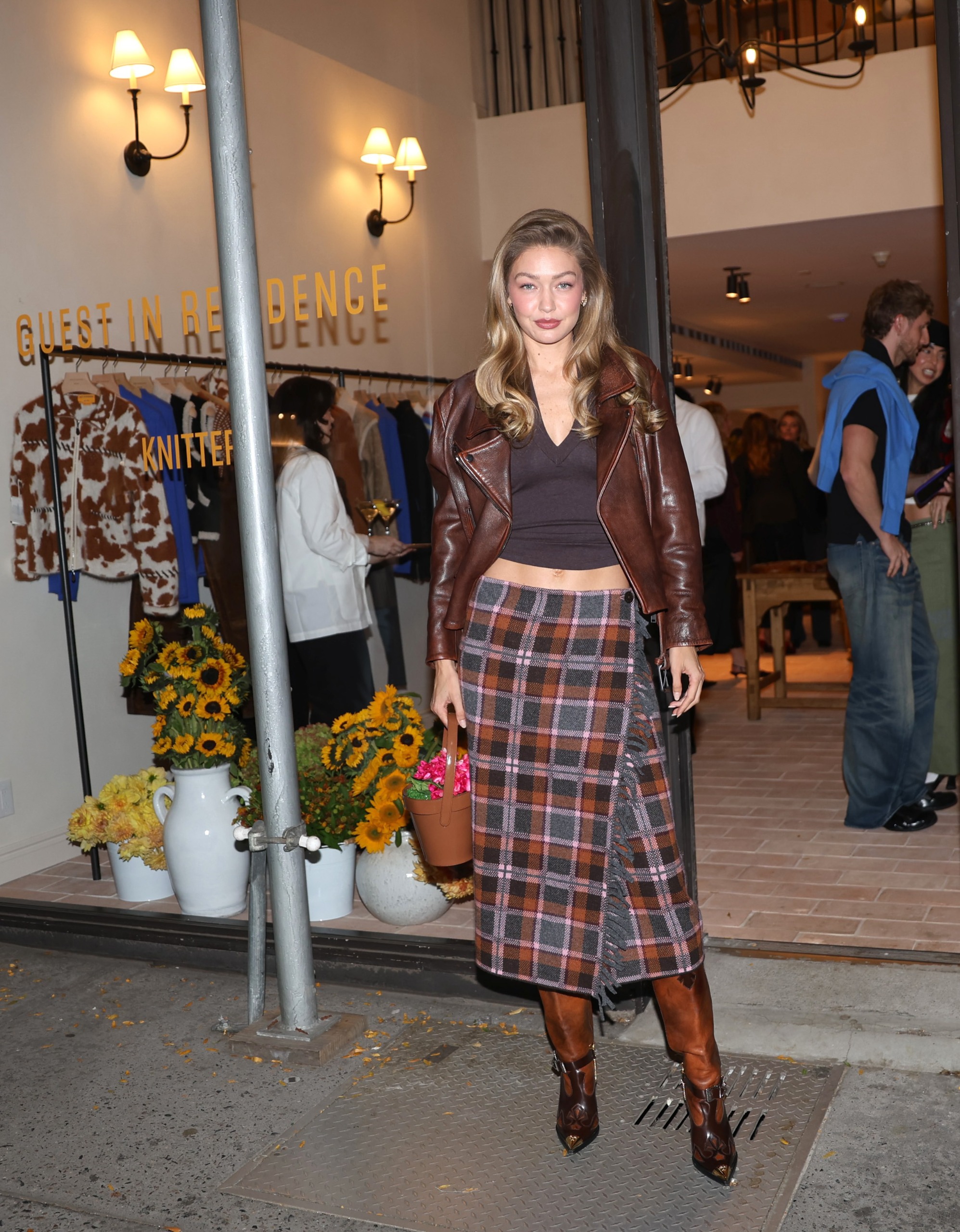 Gigi Hadid poses in front of her Guest in Residence shop wearing a wool tartan skirt with brown boots, a brown leather jacket and a v-neck top.