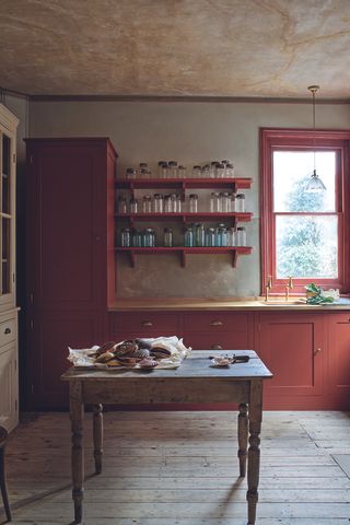 A rustic kitchen with brick red cabinets and matching window trim with wooden worktops and a red farmhouse table