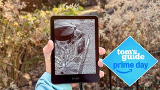 a person holding the kindle paperwhite 2024 showing a book on the e-reader with bushes in the background and a tom's guide prime day deal badge
