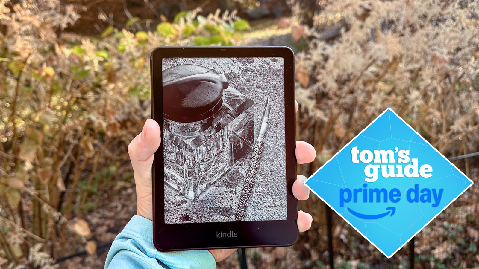 a person holding the kindle paperwhite 2024 showing a book on the e-reader with bushes in the background and a tom's guide prime day deal badge