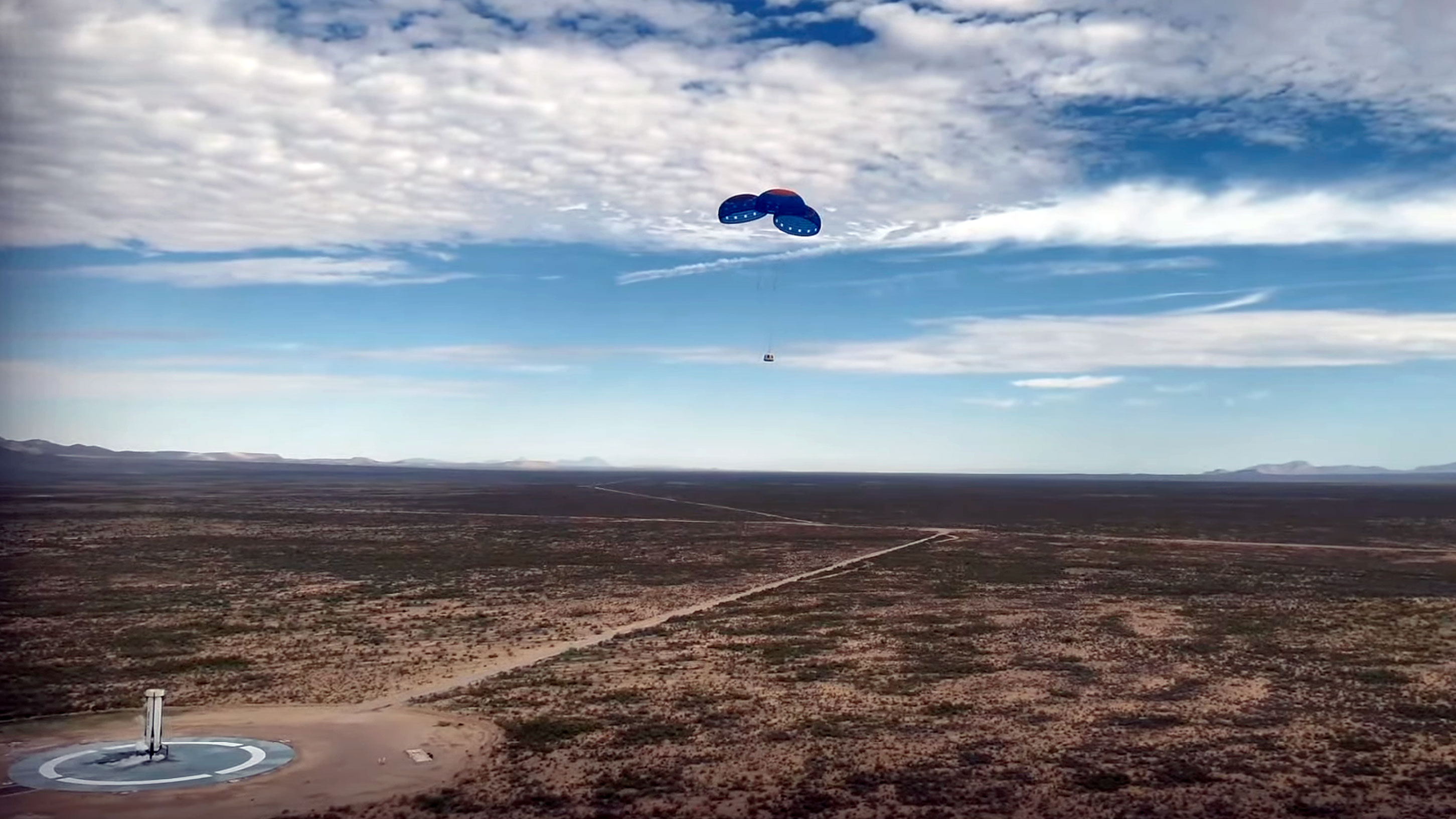 a space capsule returns to Earth under three blue and red parachutes with its reusable rocket standing on its own landing pad nearby