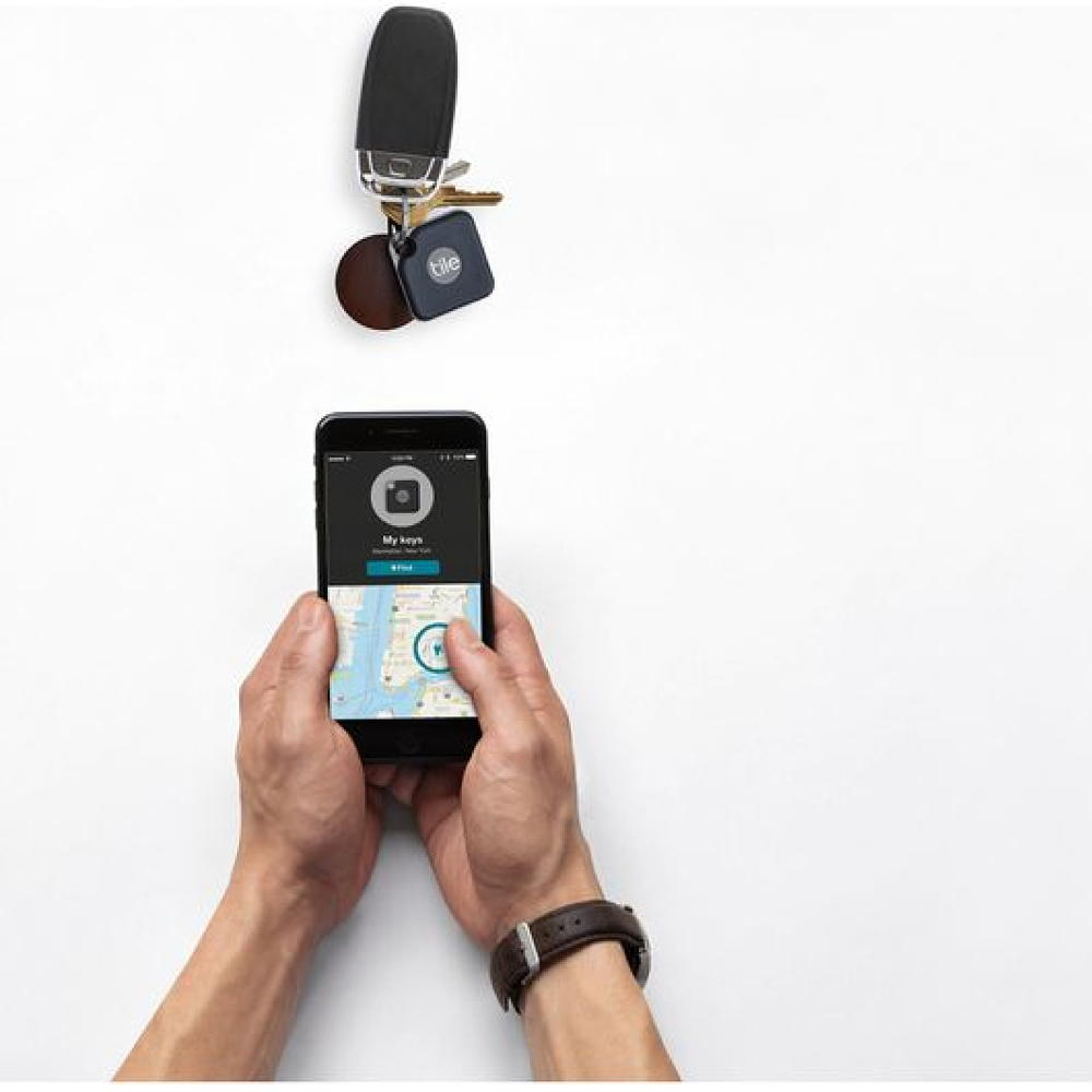 First person view of hands holding a mobile phone showing the Tile tracking app interface. A bunch of keys with a tile tracking tag sit on a surface just in front of the phone.
