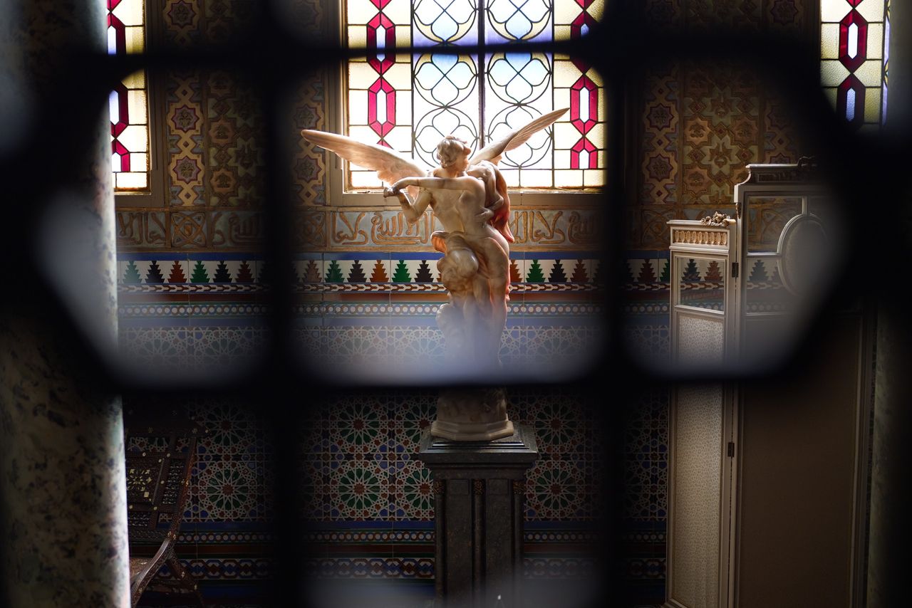 Decorative sculpture of a winged figure inside an ornate tiled interior, framed through a dark foreground railing.