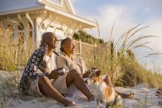 An older couple relaxes on the beach with their dog. They are drinking coffee and seem to be sitting in front of their beach house.