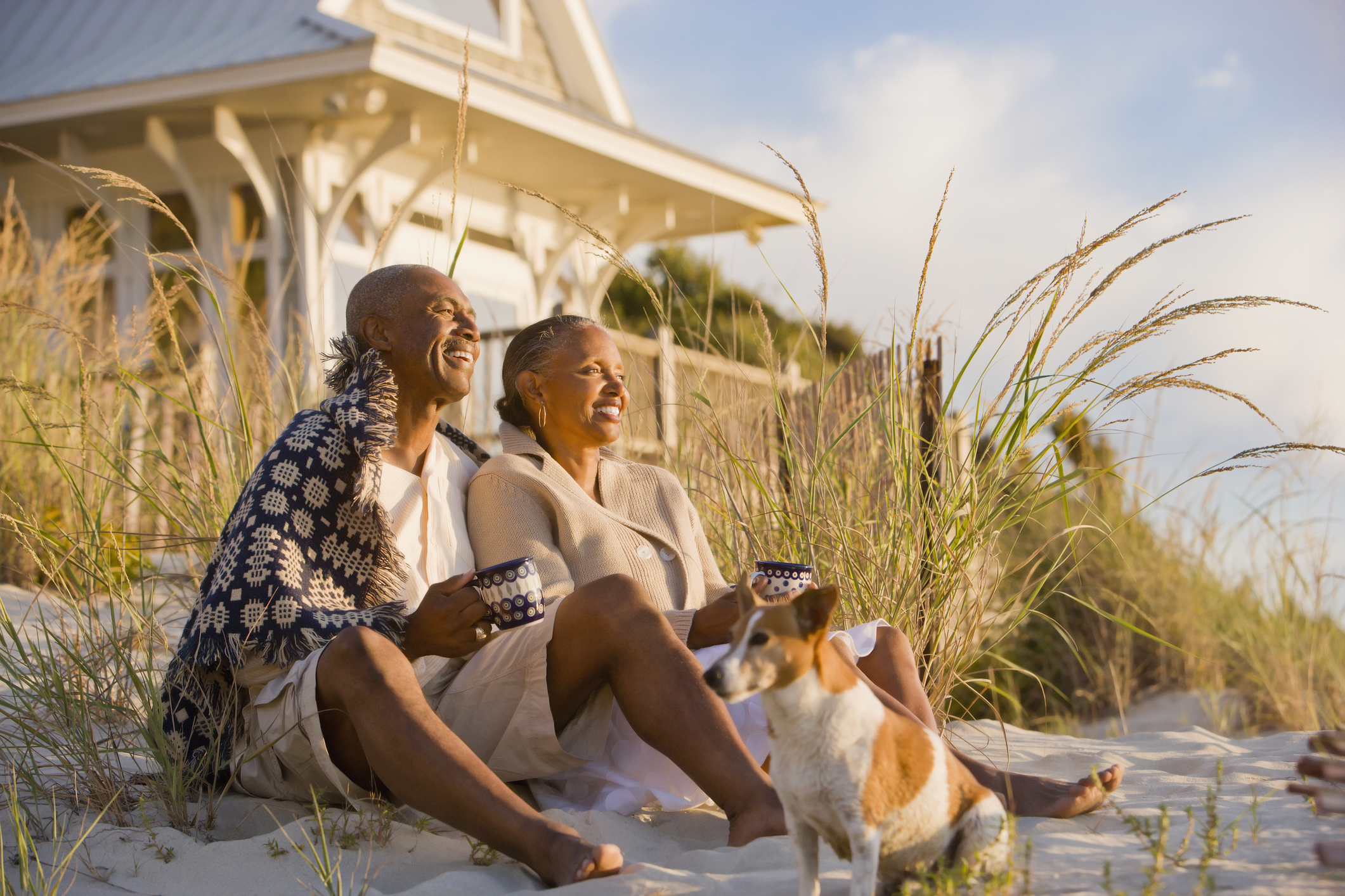 An older couple relaxes on the beach with their dog. They are drinking coffee and seem to be sitting in front of their beach house.
