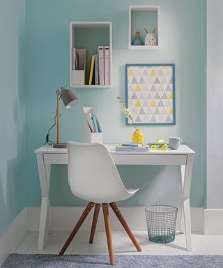 A white desk and white Eames style chair with a pale aqua wall with box shelves on the wall.