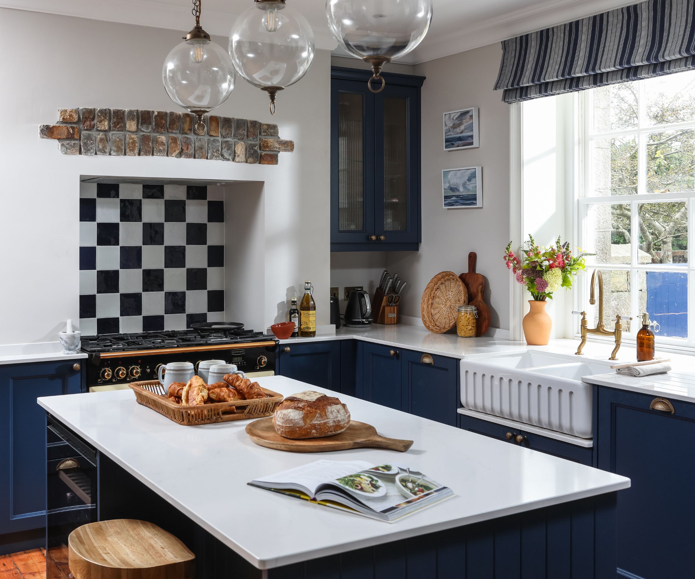 A bright country kitchen with deep navy cabinets and brass handles surrounds a white island topped with bread and pastries. A black range cooker sits beneath a brick arch and chequered tiles. A large Belfast sink and brass tap sit by a sunlit window overlooking trees.