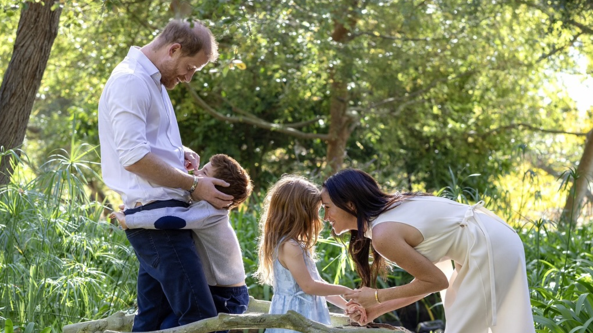 Prince Harry hugging Prince Archie and Princess Lilibet holding hands with Meghan Markle on a bridge over a stream
