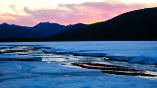 The icy landscape in Chukotka, Russia. We see ice-covered water with a mountain range in the back at sunset.