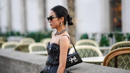 Yoyo Cao wears a black shoulder bag, black sunglasses, silver earrings, a silver large chain necklace, a black shiny leather heart-neck / shoulder-off zipper corset top, black suit pants, outside Cecilie Bahnsen, during Paris Fashion Week - Womenswear Fall Winter 2023 2024, on March 01, 2023 in Paris, France.