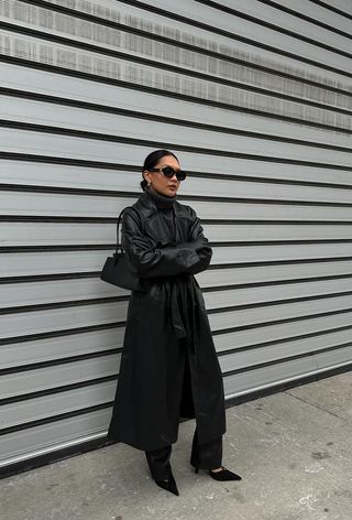 A cute work winter outfit is shown in a photo of a woman standing on the sidewalk wearing a gray turtleneck sweater with leather trousers, a leather trench coat, black pumps, and a black handbag