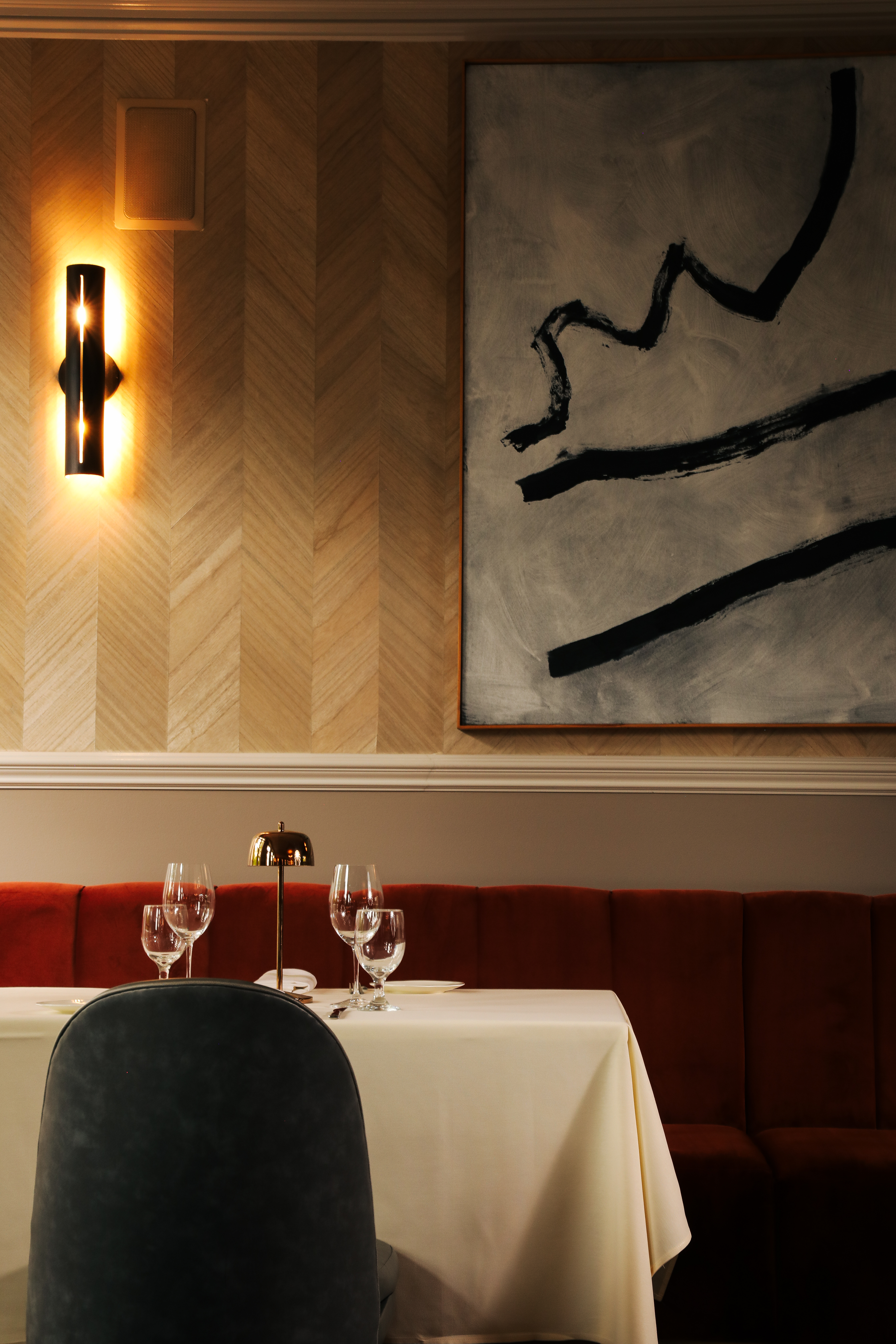 A sophisticated restaurant boasts wood-paneled walls covered in abstract artworks, softly glowing brass, cilindrical sconces, red velvet banquette seating, and green chairs, along with traditional tableware.