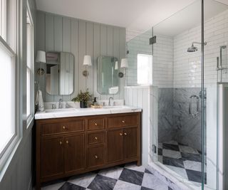 A walk-in shower in a small bathroom with marble tiles on the lower half of the wall and white subway tiles on the upper half of the wall. On the floor of the entire bathroom are black and white checkerboard tiles and a wooden double vanity with a marble countertop sits against the back wall