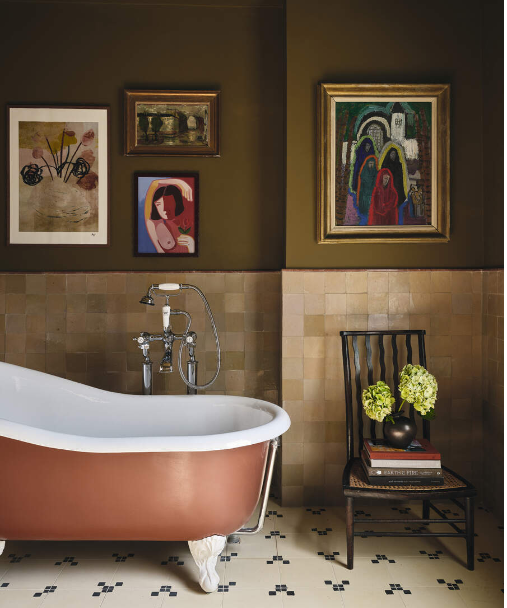Cheerful olive bathroom featuring a tiled backsplash, terracotta clawfoot tub, wall art, and a vintage wooden chair topped with books and a vase of hydrangeas