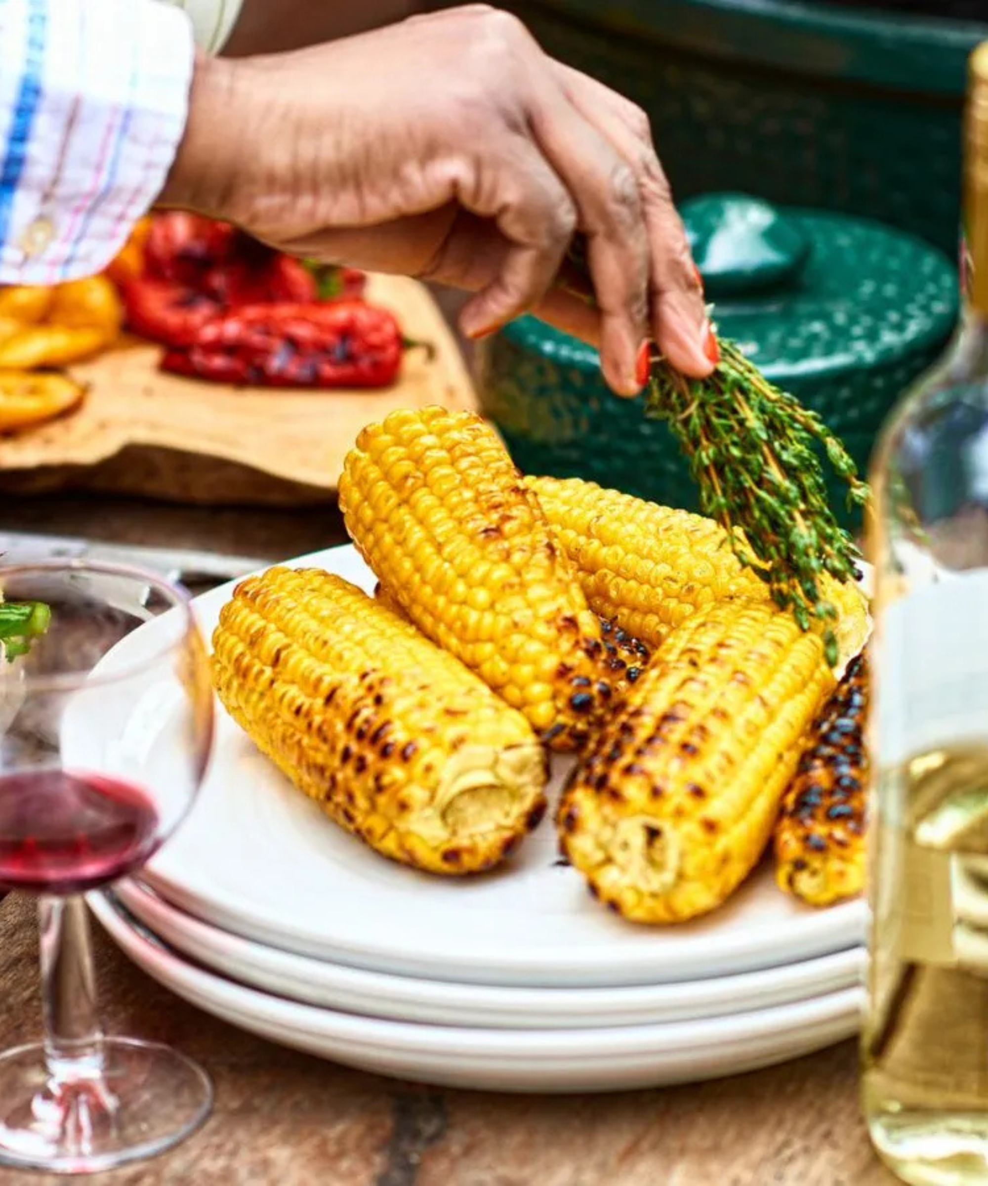 Grilled corn on a plate at a party with a glass of wine, a bottle of wine, being brushed with rosemary