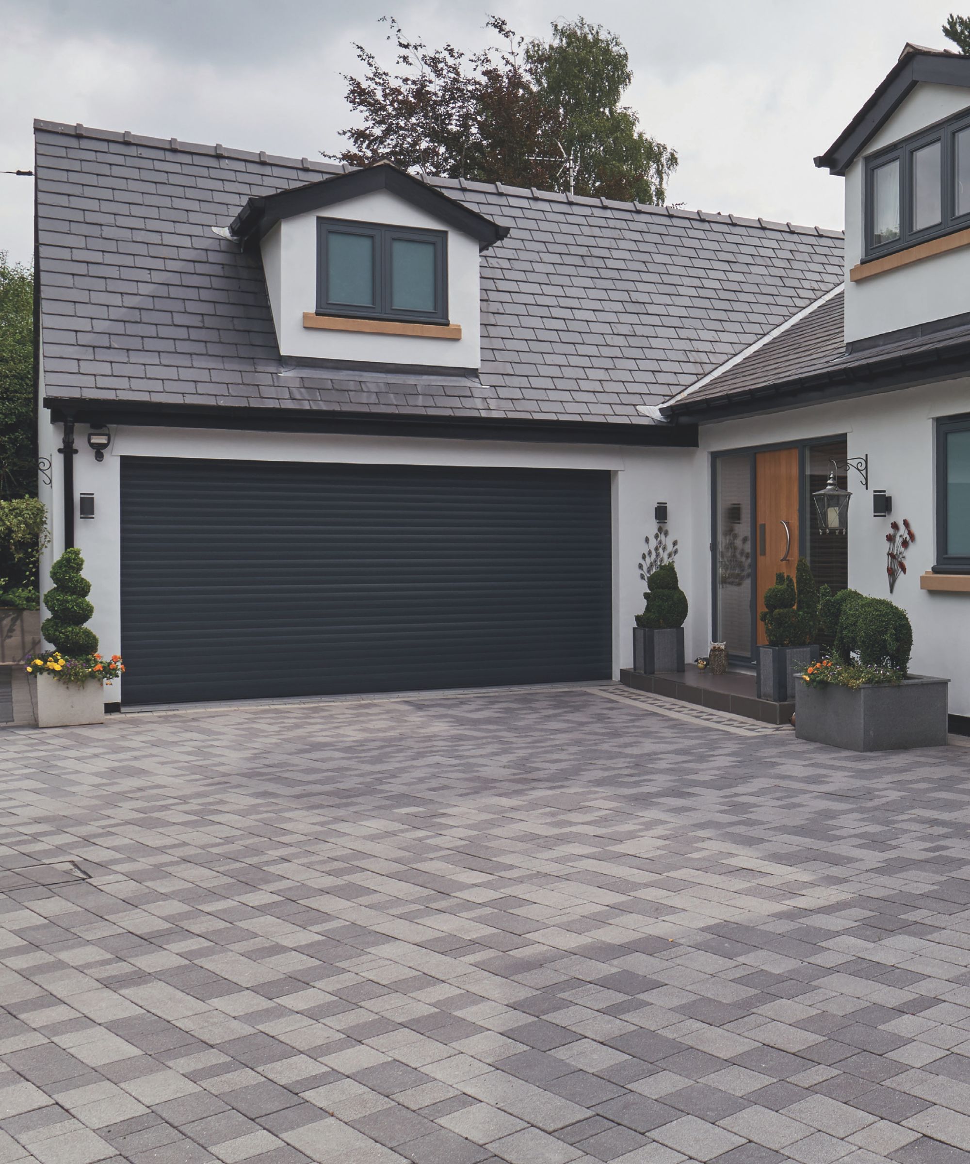Contemporary home with a slate tiled roof and white exterior, featuring a sleek black roller garage door. The driveway is laid with multi-tone grey block paving in a checkered pattern, bordered by neat planters and topiary.