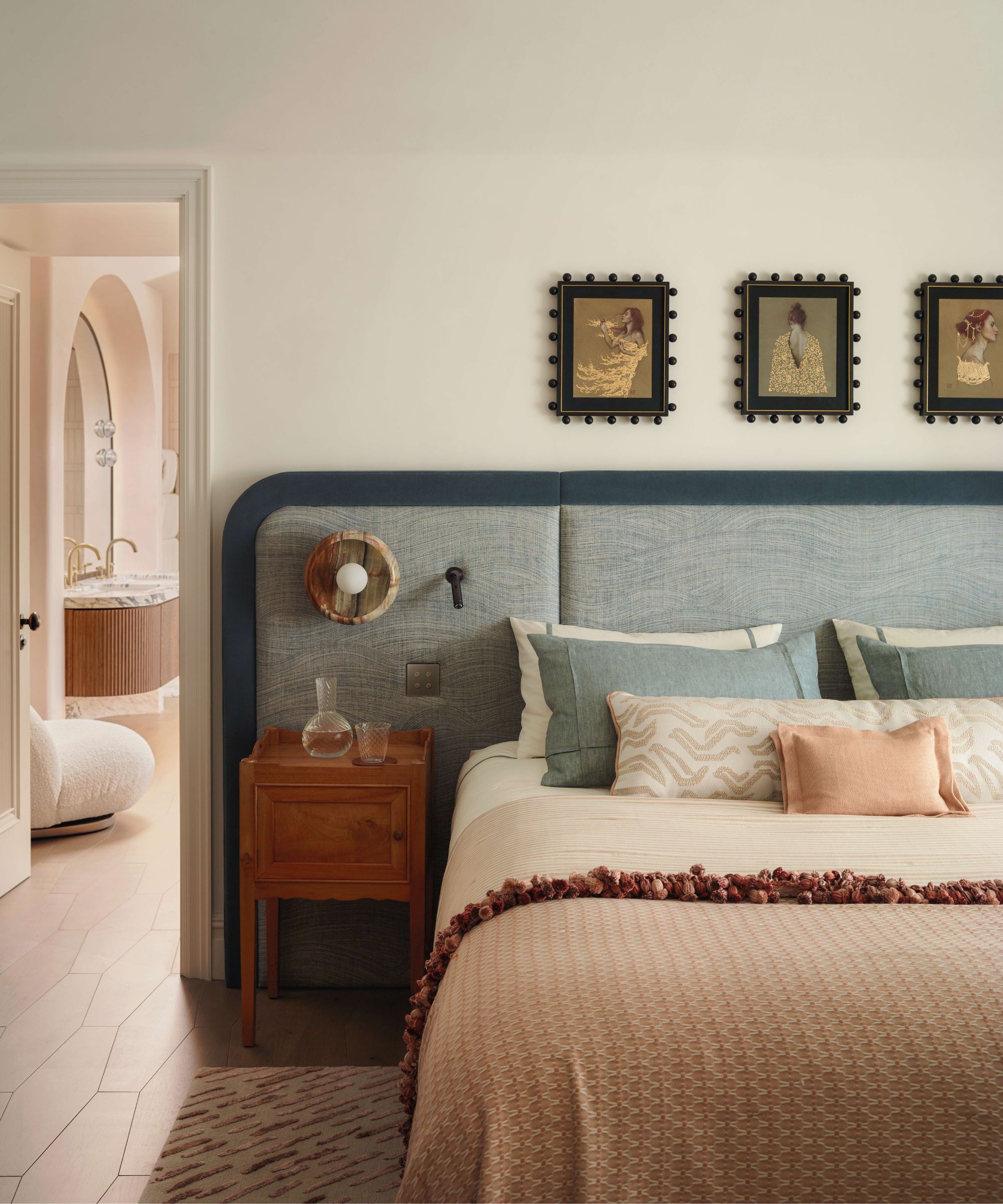 a neutral bedroom with a large blue upholstered headboard, wooden nightstand, and a soft pink and blue layered bed