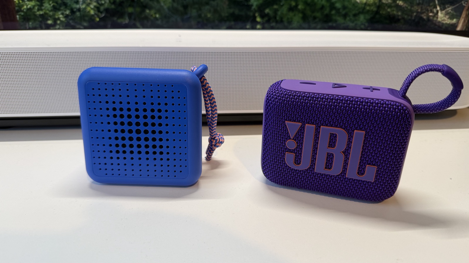 A small, Blue Ikea speaker, and a small, purple JBL speaker, photographed side-by-side on a white desk