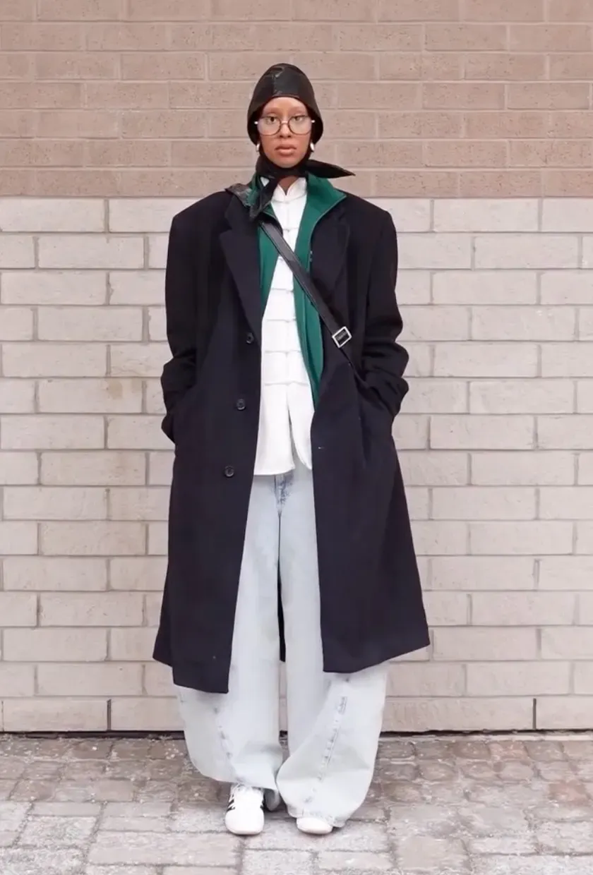 A woman wears a black jacket, white button down, barrel-leg jeans, and Adidas sneakers.