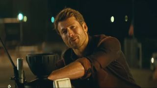 Glen Powell smiles while working at a desk with equipment at night in Twisters.