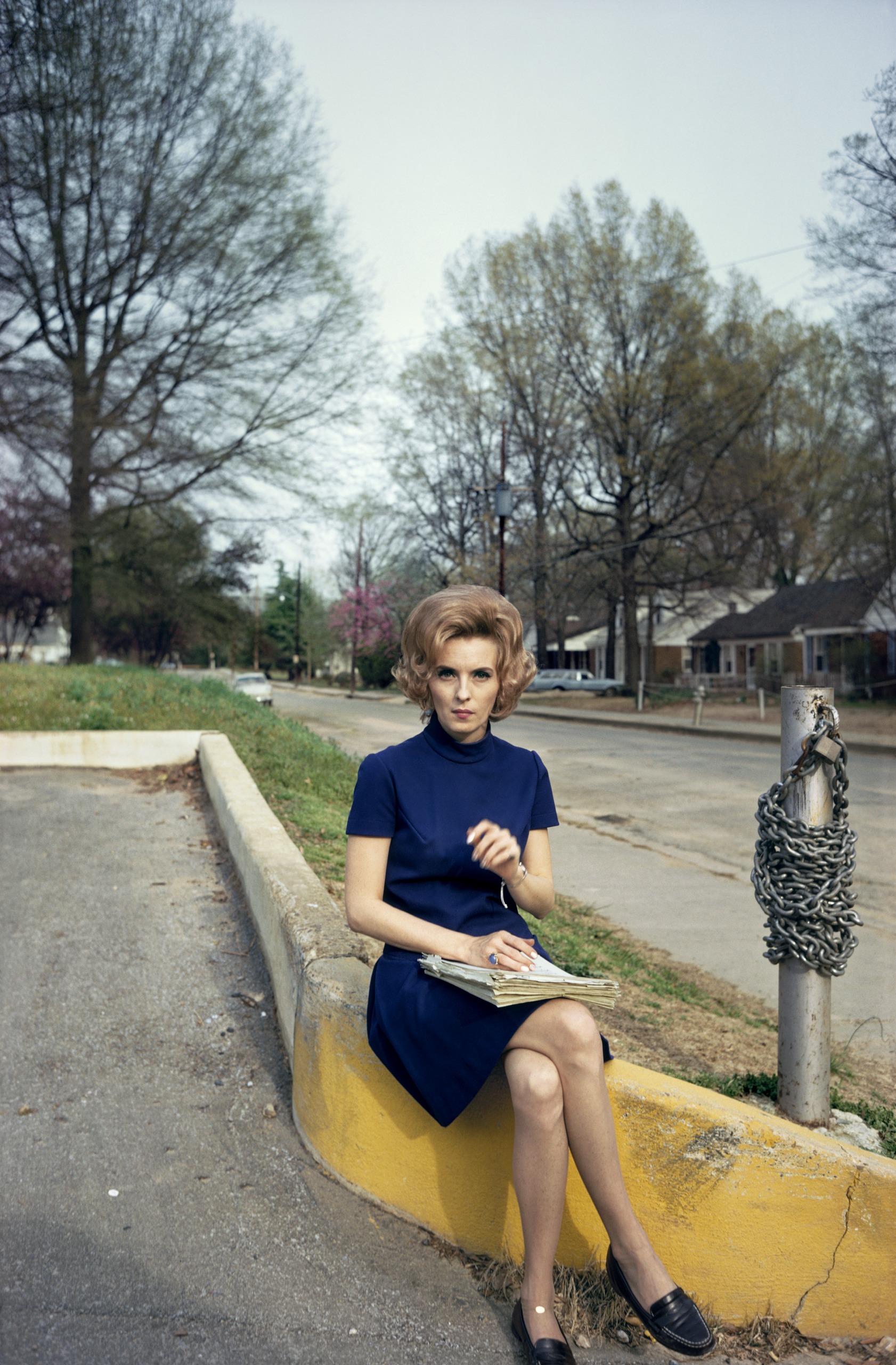 A color photograph of a woman with voluminous hair sitting on a yellow curb beside a chained post, holding a stack of papers.