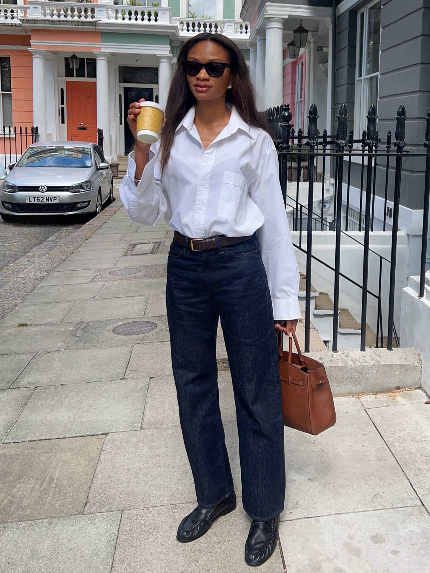 British style influencer Lorna Humphrey of Symphony of Silk poses on a London sidewalk wearing a white button-down shirt, belt, dark-wash jeans, tan top-handle bag, and black ruched loafers