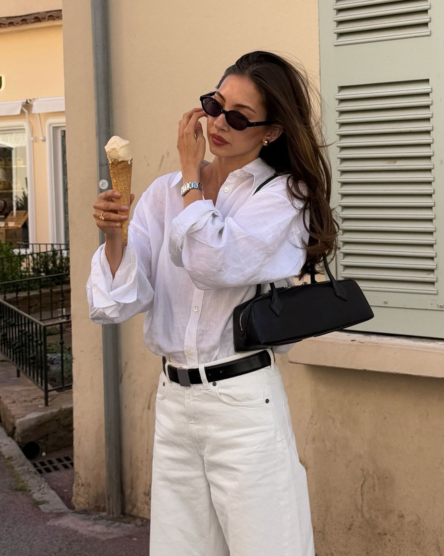 Style influencer Felicia Akerstrom poses with an ice cream cone in Saint-Tropez wearing black sunglasses on her head, a white button-down shirt, a black Alaia Le Teckel shoulder bag, black belt and ivory jeans.