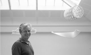 Black and white image of Hugh Dutton smiling, white walls and glass panel ceiling, white exhibition pieces hung from the ceiling