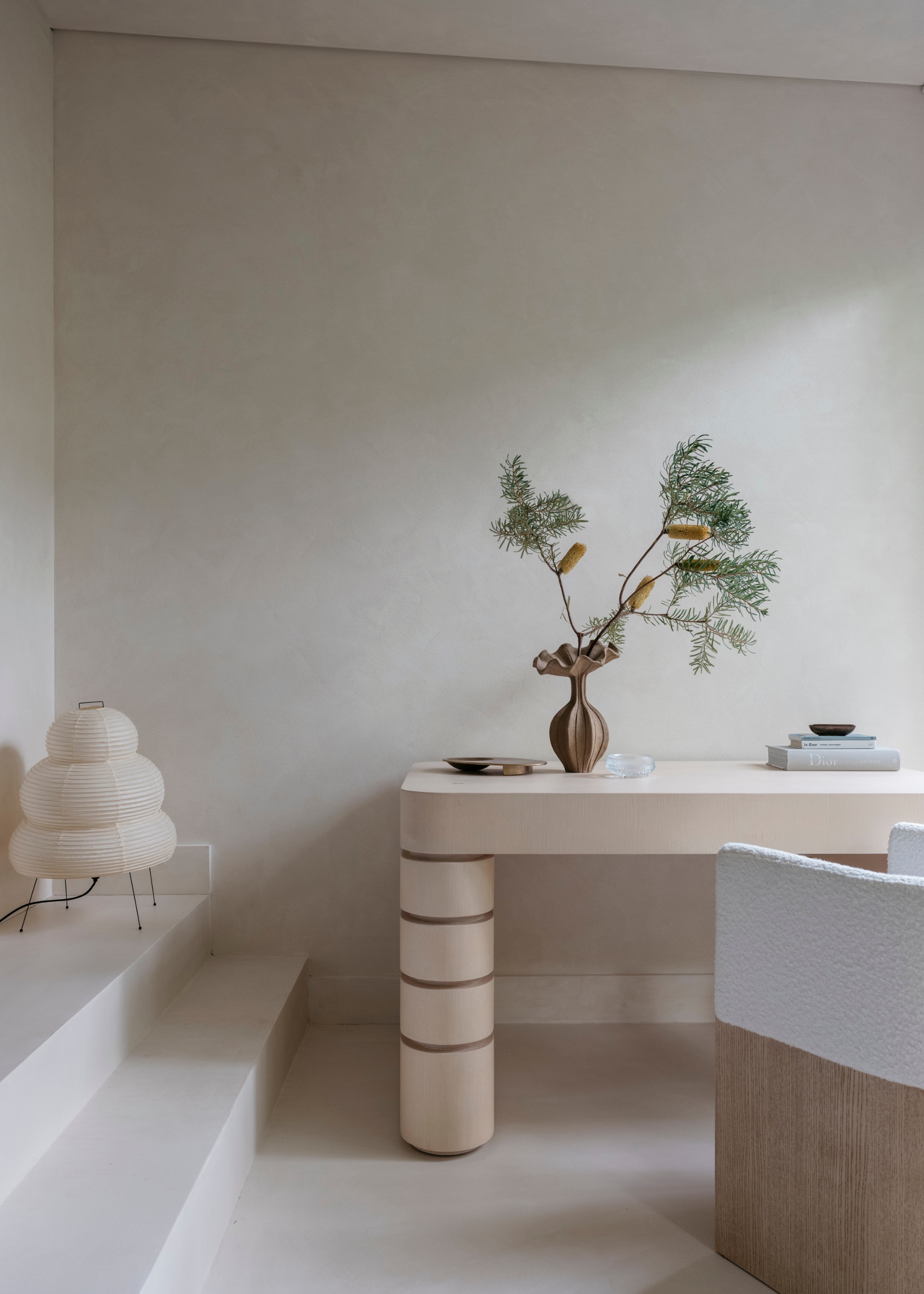 A study with a rice paper lantern, a vase of stems, a wood table and a sofa chair