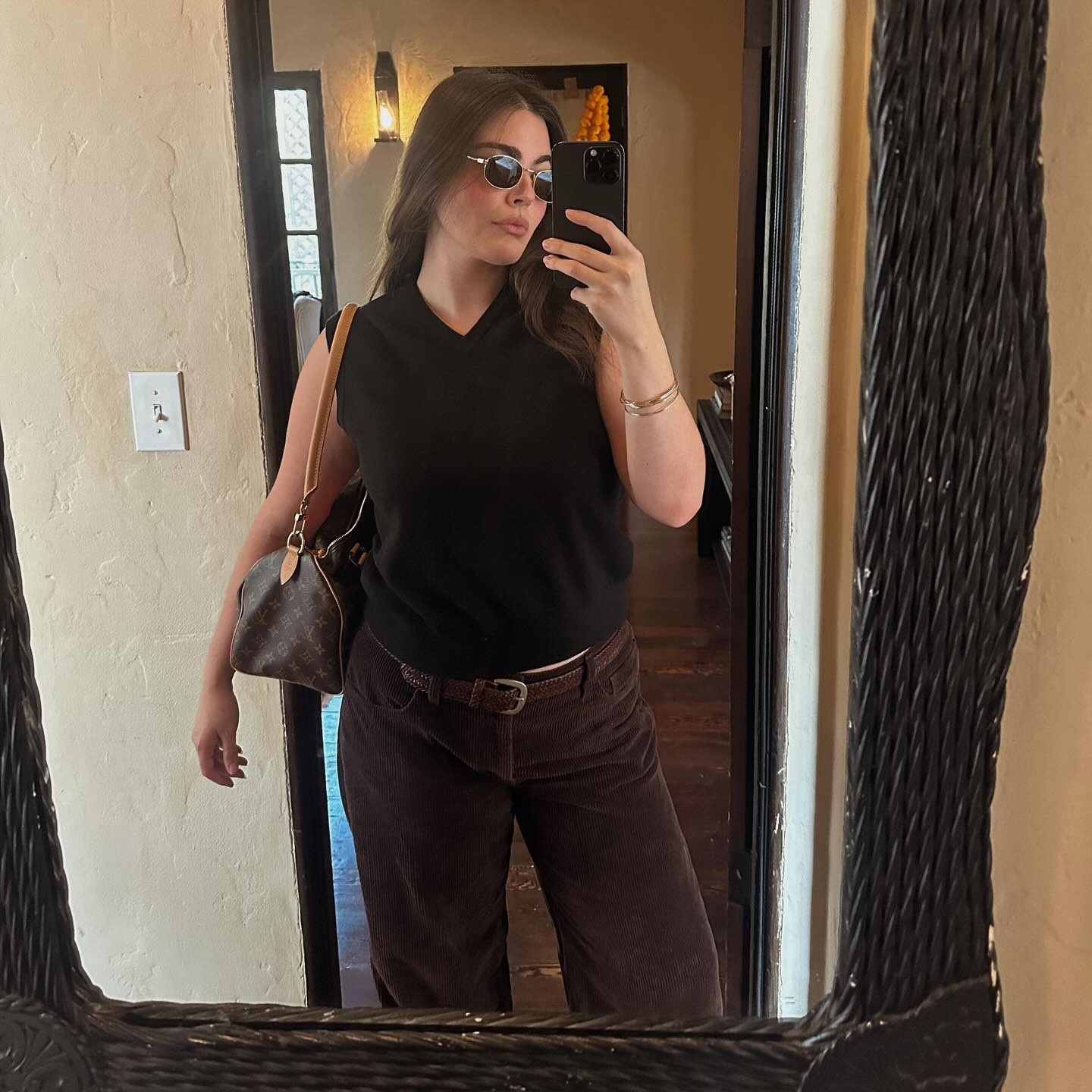 Woman wears v-neck black sweater vest with brown corduroys, a brown belt and round sunglasses.