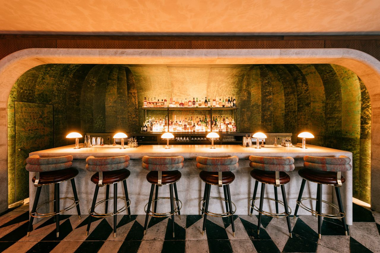 The cocktail lounge of a luxury hotel has an arched, moss green built-in bar area with warm table lamps and seating inspired by Art Deco, as well as tiled flooring in marble in black and white tones.